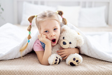 a yawning little child a blonde girl holds a teddy bear in her hands while lying under a blanket on a white bed in a bright bedroom