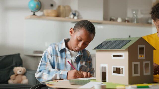 Younger brother is deeply focused on painting parts of model solar house, carefully adding color and detail to structure. His concentrated expression reflects hands-on nature of school project