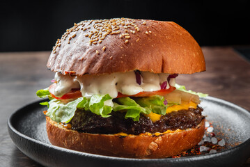 A delicious cheeseburger featuring a juicy beef patty, melted cheddar cheese, fresh lettuce, tomatoes, onions, and creamy sauce, all nestled in a sesame seed bun on a black plate.