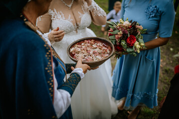 A close-up of a traditional wedding ritual, featuring a bowl of flower petals, a bride in a lace gown, and a bouquet of red and pink flowers.