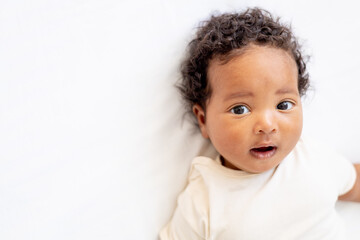 close-up portrait of a small African-American baby girl in a white bodysuit on a cotton bed at home, a six-month-old smiling joyful black baby