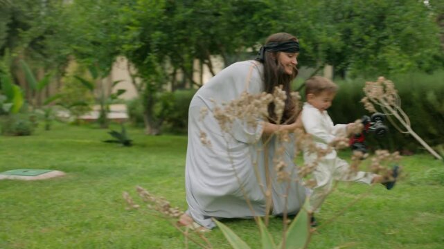 Boy joyfully runs through lush garden holding toy car, laughing as his blindfolded mother playfully chases him. She eventually catches him, lifting him up and planting a loving kiss on his cheek