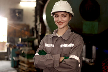 Fototapeta premium Portrait of industrial engineer woman with protective workwear and safely helmet stands with arms crossed at manufacturing factory. Smiling beautiful female worker work at steelmaking metal industry