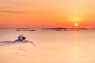 Lobster boat heading out to sea with full, golden sun just over the horizon. Islands on the horizon. Mid-coast Maine.