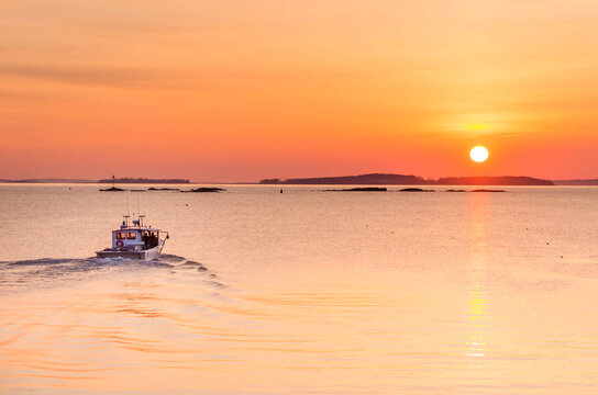 Lobster boat heading out to sea with full, golden sun just over the horizon. Islands on the horizon. Mid-coast Maine.