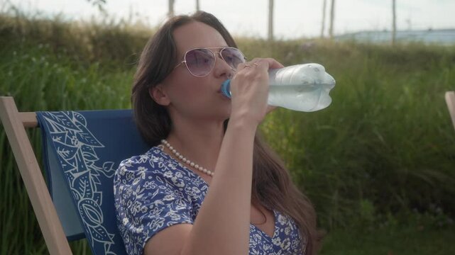 A young woman sits on a chaise longue on a hot summer day, drinking cold still water from a plastic bottle with an eco-friendly tethered cap, complying with EU regulations to reduce plastic waste.