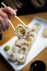 Close-up of a person holding a sushi roll with chopsticks with sushi set in the background