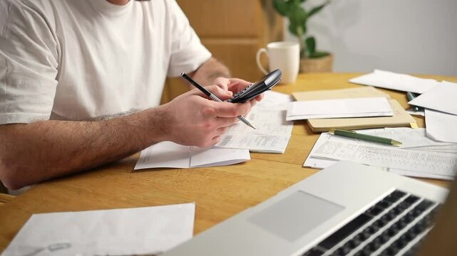 A man is sitting in a kitchen checking bills on a computer. The table is cluttered with a calculator, papers, invoices, and letters, emphasizing financial tasks and household management.