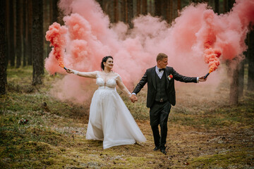 Bride and groom holding pink smoke bombs while walking through a forest, creating a unique,...