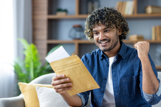 Successful and joyful man sitting on sofa in living room, holding postal envelope letter in hands, celebrating victory and successful results obtained in notified .
