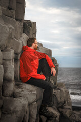 A male adult hiker in a red jacket sits on hexagonal basalt columns near Hálsanefshellir Cave,...