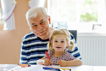 Cute little baby toddler girl and handsome senior grandfather painting with colorful pencils at...