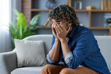 Desperate and depressed man sitting on sofa in living room at home, burying face in hands, feeling...