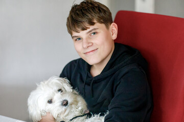 Happy teenager boy and cute maltese puppy together. Handsome child holding his dog at home, hugging. Pet love, family dog.