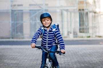 Obraz premium Schoolkid boy in safety helmet riding with bike in the city with backpack. Happy child in colorful clothes biking on bicycle on way to school. Safe way for kids outdoors to school
