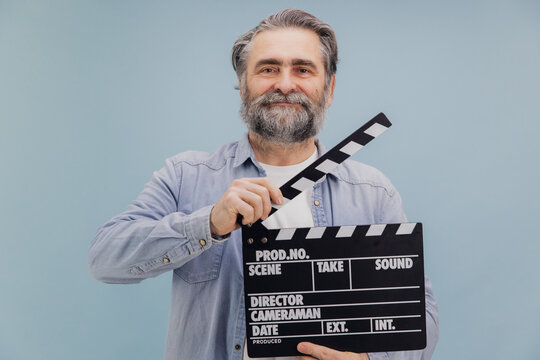 Senior film director holding movie clapper board smiling on light blue background