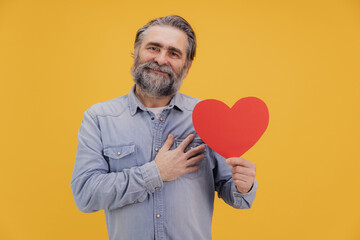 Senior man showing red heart and expressing love on yellow background