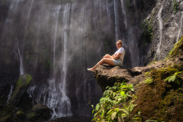 A tranquil scene showcases a woman peacefully sitting beside a magnificent, cascading Tumpak Sewu waterfall, enveloped by lush greenery and a gentle, refreshing mist in the air, East Java, Indonesia