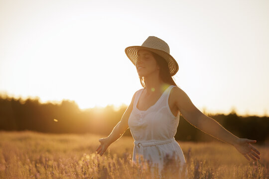 Young woman enjoys and breathes the pure air and the aroma of the lavender field. Woman with a straw hat and white dress, has her arms open enjoying the moment with the sunset in the background.