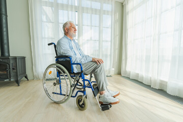 Sad senior man sitting in wheelchair looking out window in retirement home hospital. Old paralyzed...