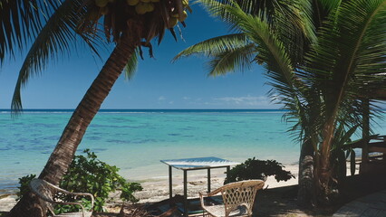 Tranquil beach scene showcasing swaying palm trees, a cozy table and chairs, and stunning turquoise waters, creating an inviting atmosphere for relaxation and enjoyment under the sun