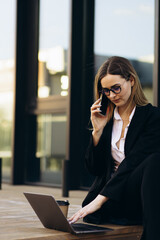 Business woman working on laptop outside the building and drinking coffee