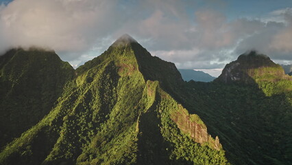 Fototapeta premium Aerial view panorama of dramatic mountain range ridge peaks covered green hill jungle forest under sunset clouds sky. Moorea tropical island, French Polynesia. Remote wild nature travel. Drone flight