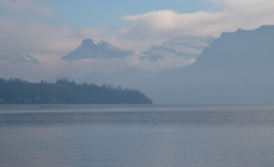 Fog over the lake and river surrounded with high mountains with snow on tops in sunny day with blue sky