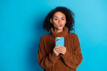 Young woman holding smartphone against blue background wearing brown sweater making playful facial...