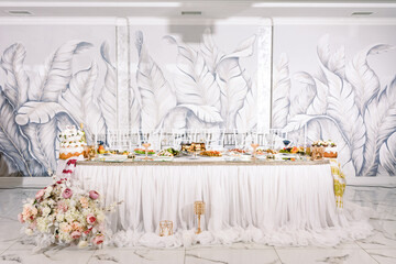 A lavishly decorated wedding reception table showcases an array of desserts, appetizers, and a beautiful floral arrangement.  The backdrop features a stunning leaf mural.