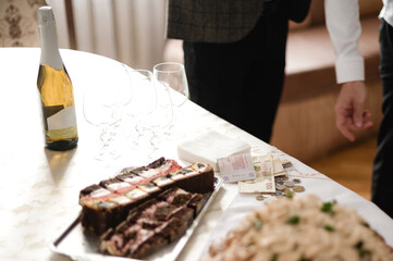 A table displays sliced layered cake, empty glasses, a bottle of sparkling wine, and some banknotes...