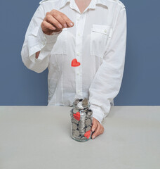 Woman in white shirt throwing red heart into transparent jar with silver coins. Concept of doing...