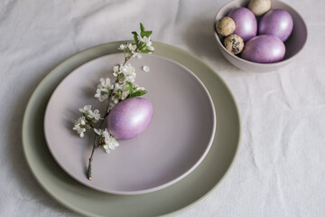 Easter table setting with minimalist floral decor. Pastel purple Easter eggs in a gray plate on linen tablecloth