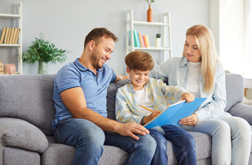 Smiling parents sitting on couch with their cheerful son between them writing in notebook. Happy mother and father spending leisure time with child, helping him with homework. Family bonds concept.