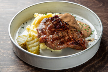 A delicious plate featuring a grilled steak served on creamy mashed potatoes, accompanied by a rich sauce and pickle slices. Perfect for food-related projects and menus.