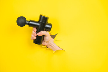 Naklejka premium Female hand sticking out with massage gun from yellow paper background. Copy space. 