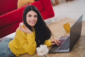 Casual young woman enjoying leisure time with laptop and marshmallows at home, wearing yellow...