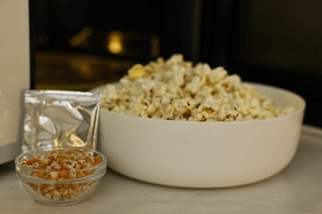 Bowl of popcorn, kernels and bag near open microwave oven on white marble table, closeup