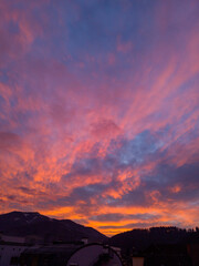 Stunning sunset paints sky with vibrant hues of red, orange, and pink, blending into deep blues and purples. Silhouetted hills create contrast against fiery horizon, adding drama to serene landscape.