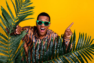 Stylish man with green sunglasses interacting with tropical leaves against a vibrant yellow...