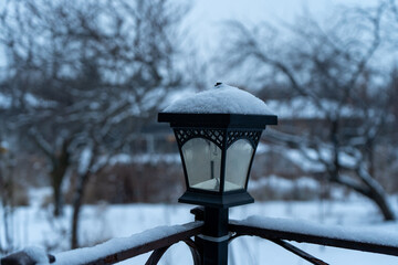 A streetlight mounted on a fence is dusted with snow, casting a soft glow on the serene, snowy landscape. This peaceful winter scene highlights the quiet beauty of a snowy evening.