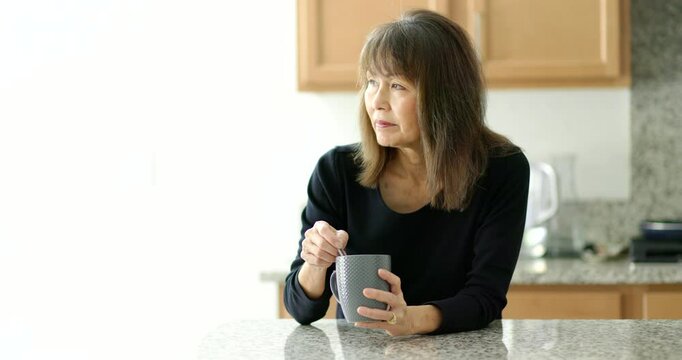 Woman relaxing stirring coffee cup standing at kitchen counter near bright window