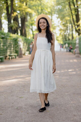 carefree African American woman in white dress and straw hat walks in park and poses against background of green hedge. daytime for relaxation. beautiful female fashion model, sunlight summer time