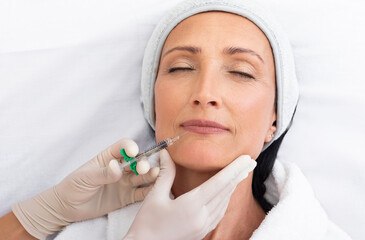 Naklejka premium Closeup shot of middle aged woman getting lip filler at clinic, doctor making hyaluronic acid injection for beautiful lady