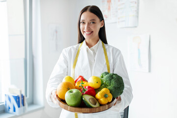 Portrait of joyful European nutritionist in lab coat holding plate of fresh fruits and veggies at...