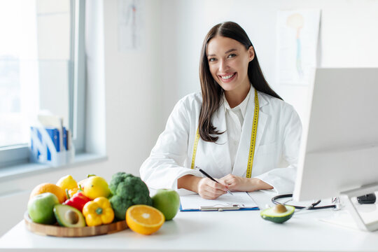 Portrait of cheerful female dietologist writing healthy eating plan or weight loss diet for patient, sitting at desk with plate of fresh fruits and vegetables