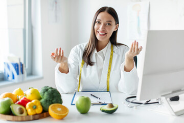Professional woman nutritionist working in office in front of computer and gesturing at camera,...