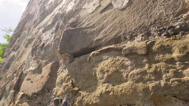 Sunlight illuminates tall stone wall, its textured surface revealing various sizes and shapes of rocks, creating sense of strength and history against backdrop of clear blue sky