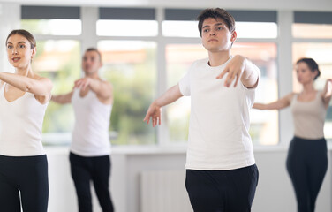 Young guy, amateur ballet dancer, dressed in white t-shirt and black sweatpants, executing dance moves with focused precision at group rehearsal in studio....