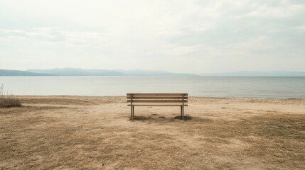 Lonely bench on sandy beach overlooking calm lake and distant mountains under a cloudy sky.